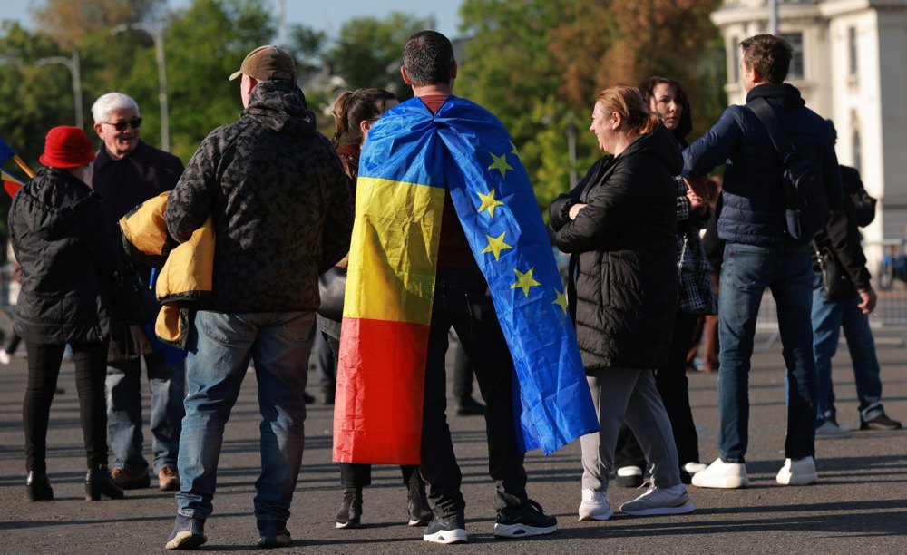 Чоловік, загорнутий у прапори Румунії та Європейського Союзу, разом з іншими активістами під час мітингу підтримки
прем'єр-міністра Боложана (голови партії <i>PSD</i>) біля штаб-квартири уряду в Бухаресті, 3 травня 2026 року